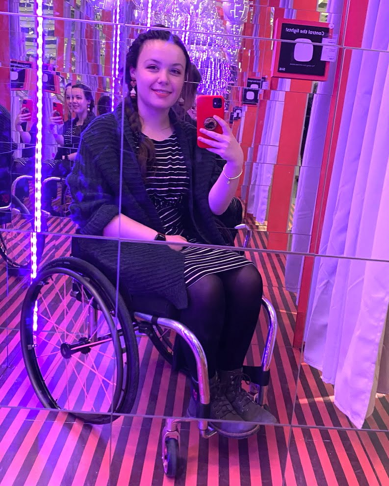 Woman in wheelchair wearing a striped dress. She is sat in a room filled with mirrors, which is reflecting purple light.