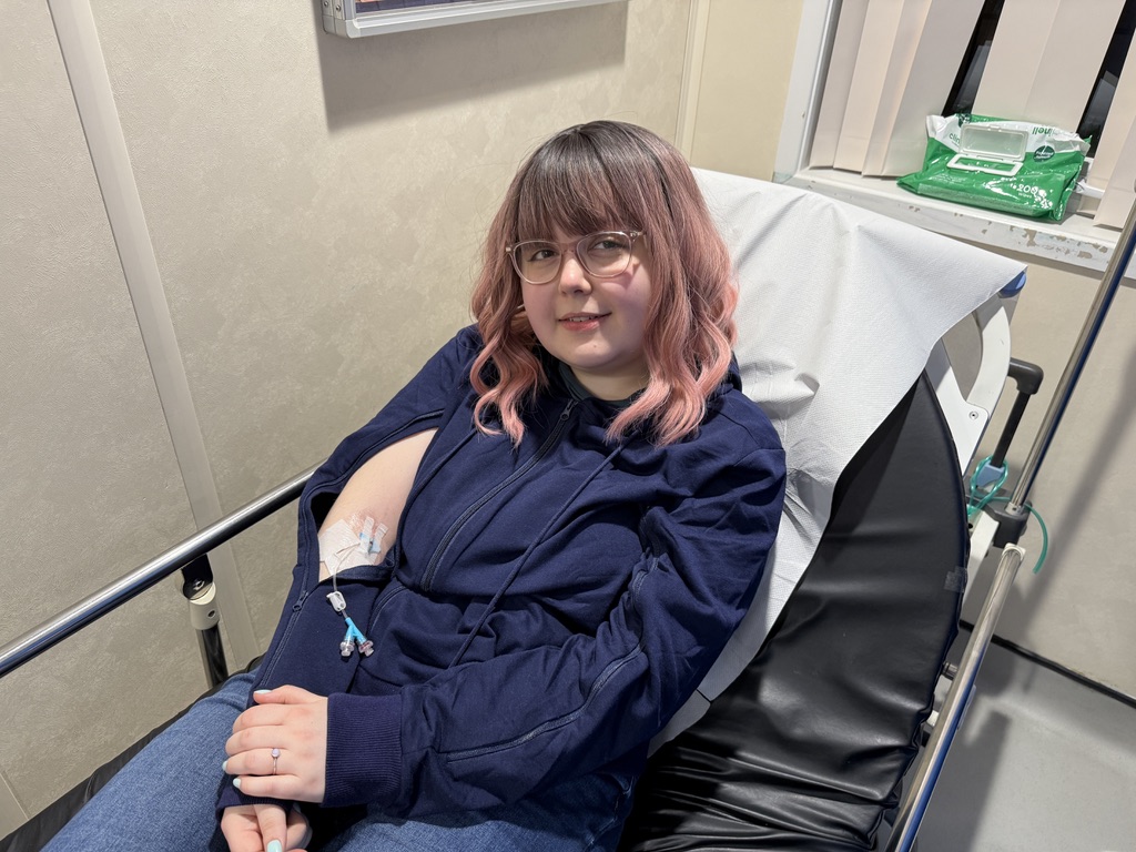 A young woman with pink hair, sitting on a hospital bed, wearing a blue hoodie with an IV access zipper. She has a cannula in her arm.