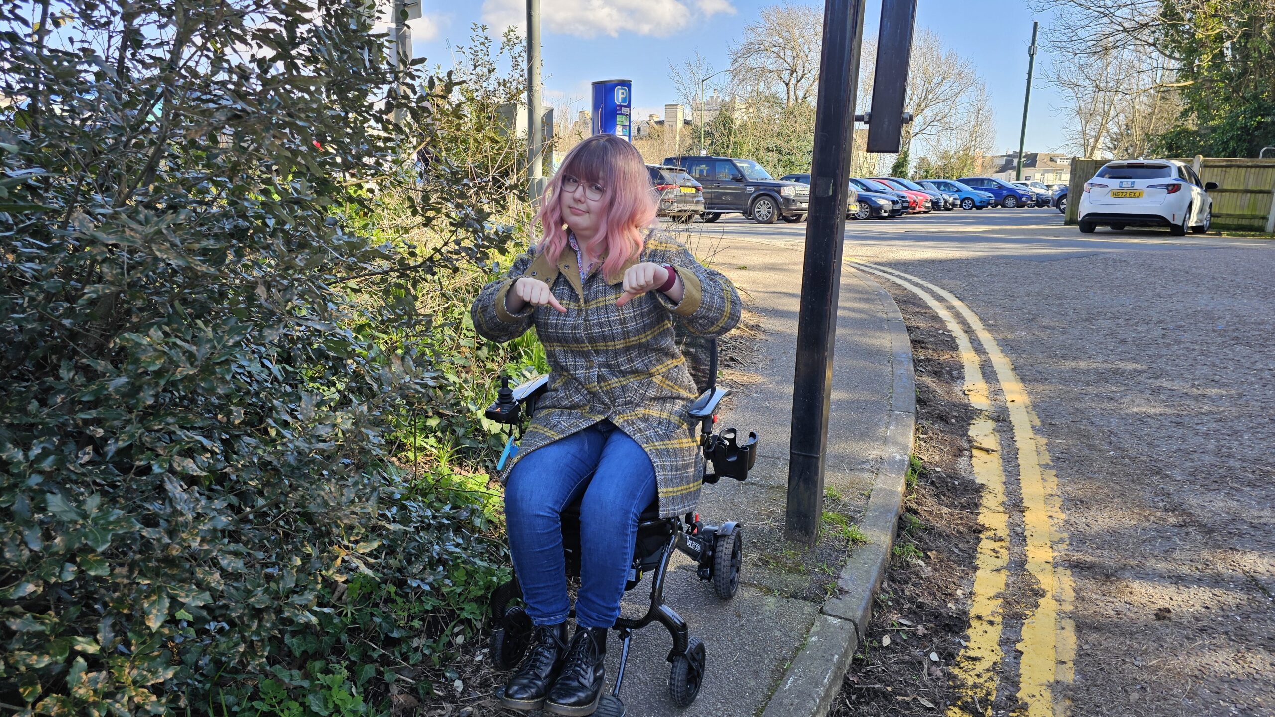 A female wheelchair user with pink hair, sitting in a wheelchair on a pavement which is blocked by weeds and debris. She is pointing her thumbs down and looking sad because there's no accessibility.