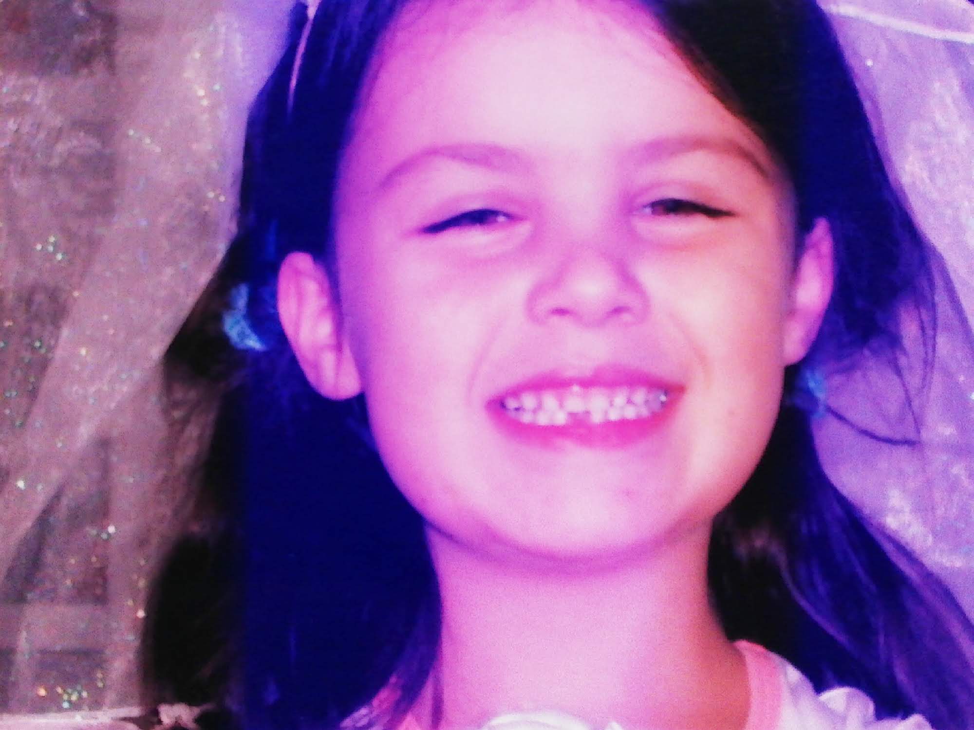 Small child with brown hair, smiling at the camera missing her front teeth.