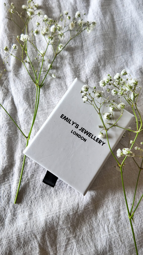 A white box with a drawer, against a linen background with babies breath scattered around. The box has the word 'Emily's Jewellery London' printed on it. 