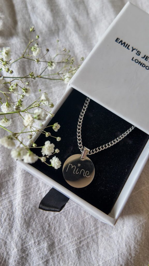 A white box, with a drawer which is half open, revealing a silver necklace inside which has a disc charm, that reads the word 'Mine' in handwritten folder. Nearby there is babies breath scattered. 