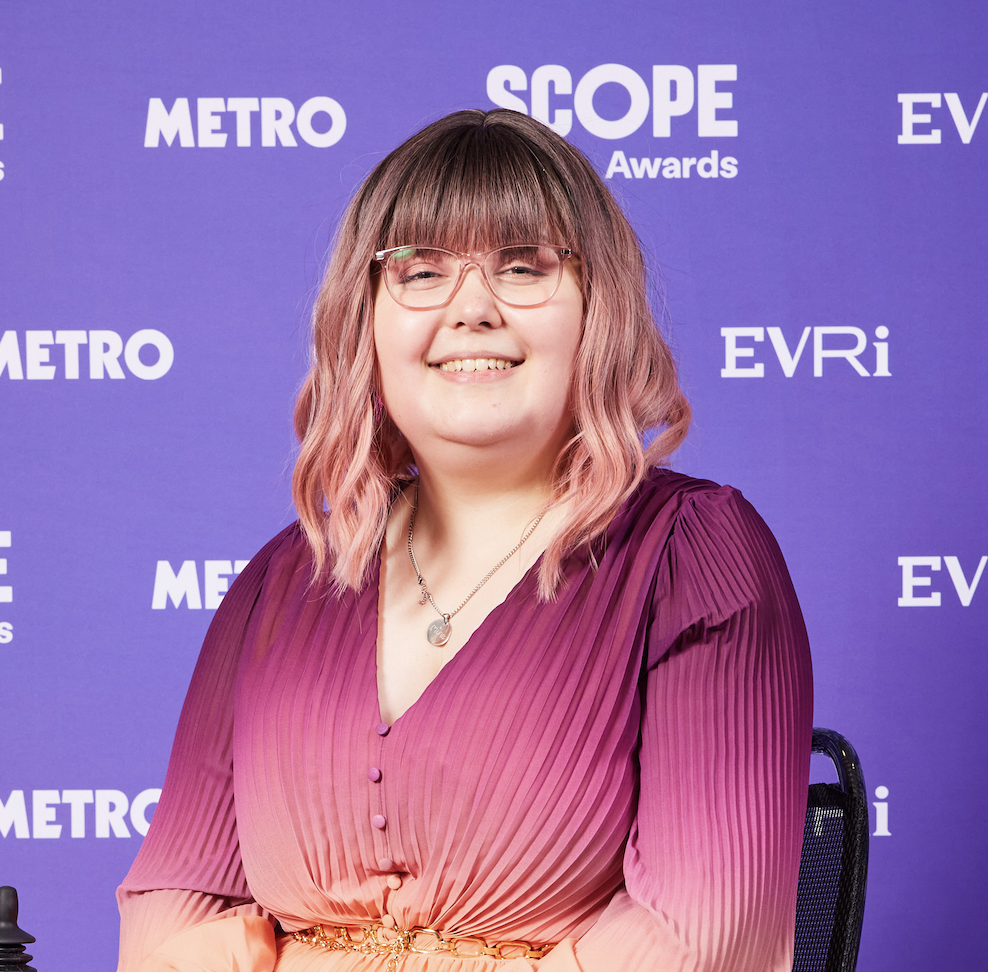 Imali, a young woman with pink ombre hair and glasses, wearing an ombre pink to orange dress. She also has a silver necklace.