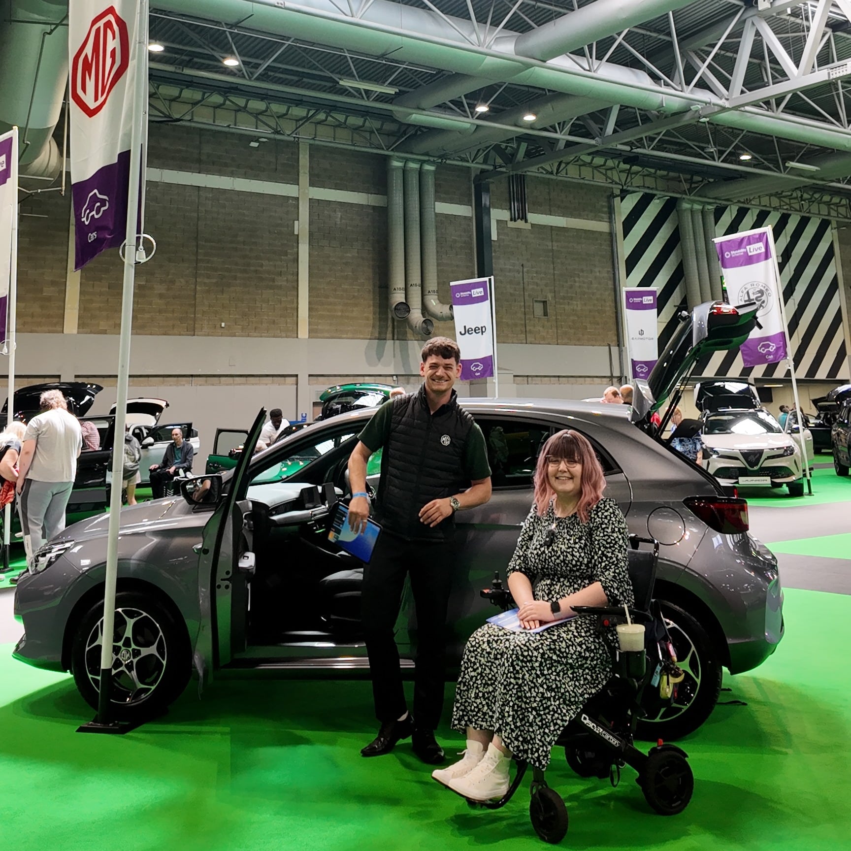 Imali, a woman with pink hair sitting in an abstract print dress in a wheelchair, next to a young white man. They are both standing next to a car.