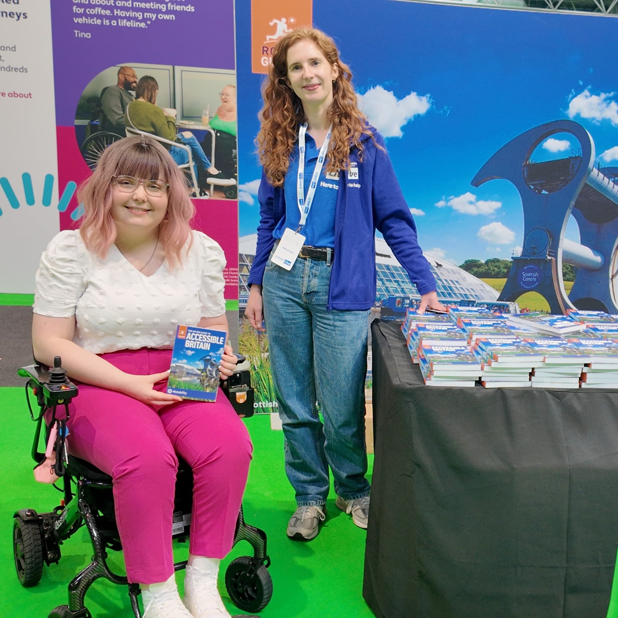 Imali, a white woman with pink hair, wearing a white top and pink trousers, is in a wheelchair next to a stand. On the stand is a white woman with red hair, wearing a blue jacket that says 'rough guides'.