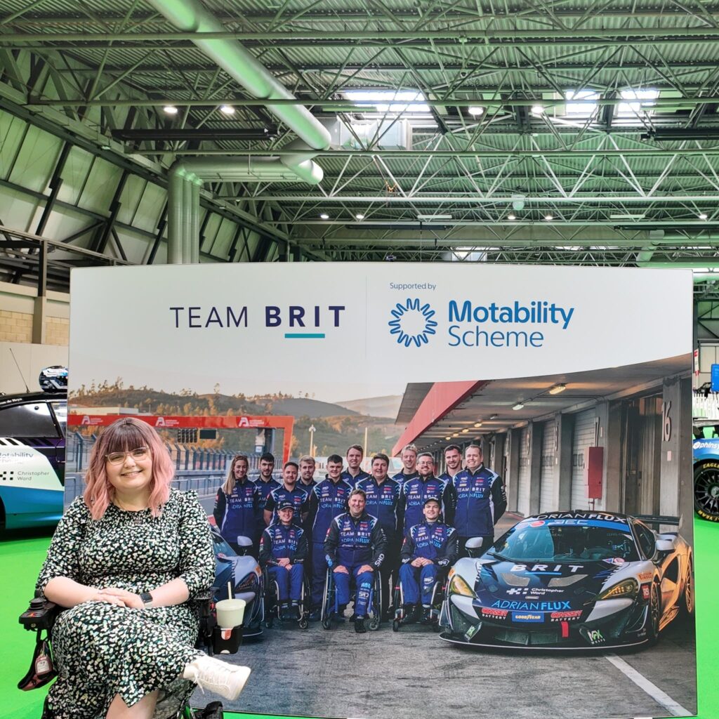 Imali, a white woman with pink hair, sitting in a wheelchair in front of a Team Brit banner.