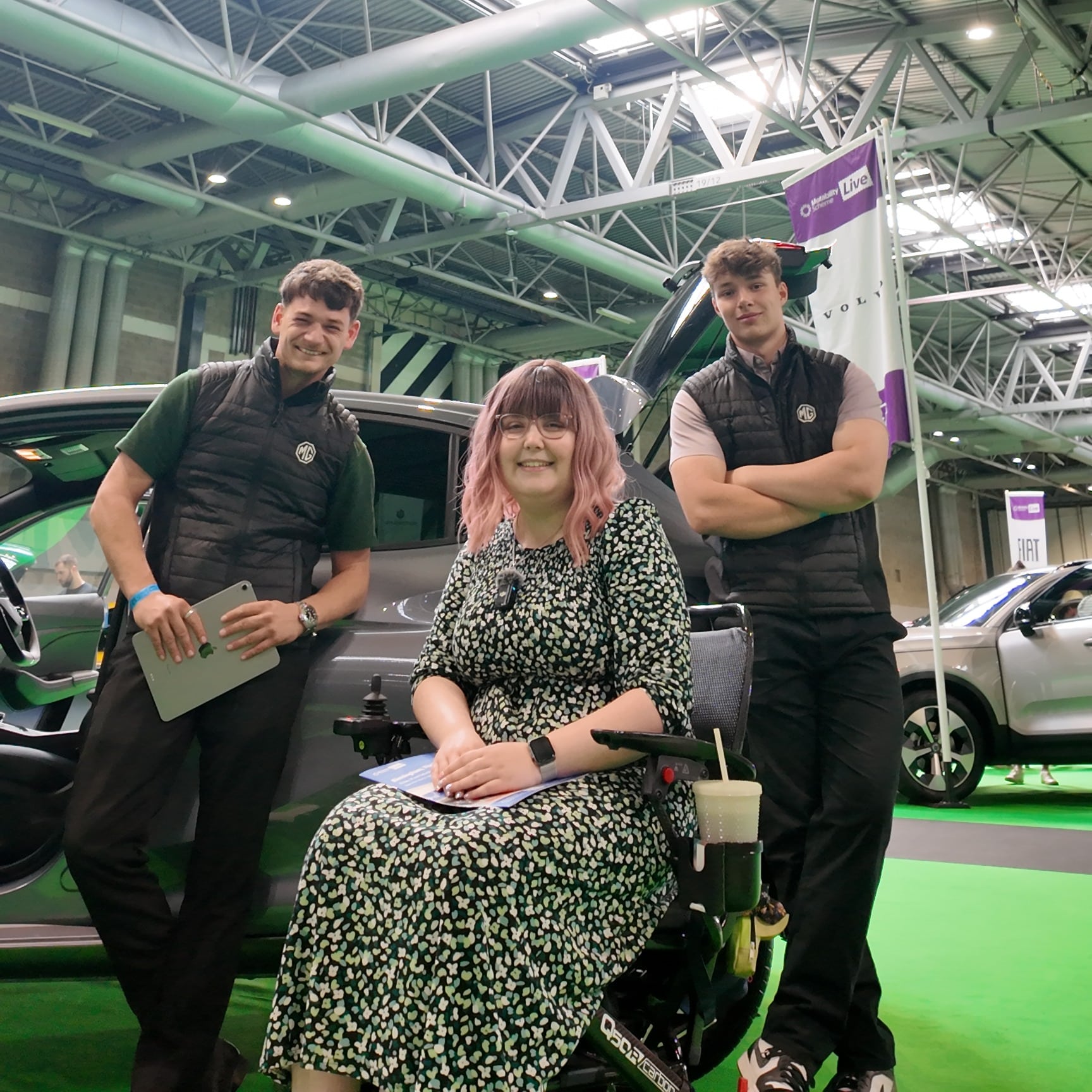 Imali, a white woman with pink hair sitting in a wheelchair, with two young men standing next to her in front of an MG car.