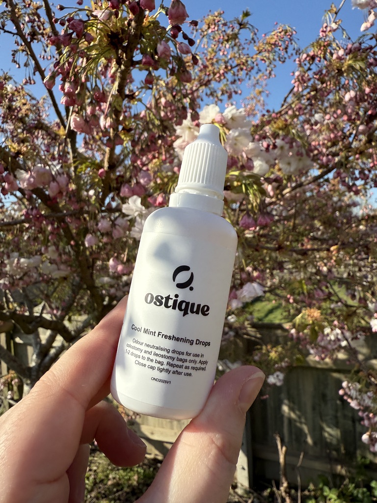 A white hand, holding a white bottle of stoma scent oil, in front of a pink blossom tree. 