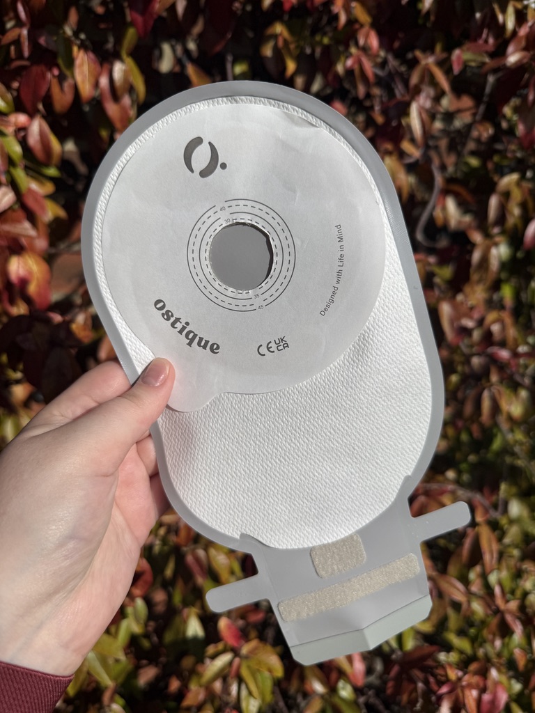 A white hand, holding a grey Ostique Onovus Solo Ostomy bag which has a white paper-like background and an adhesion patch that says Ostique.