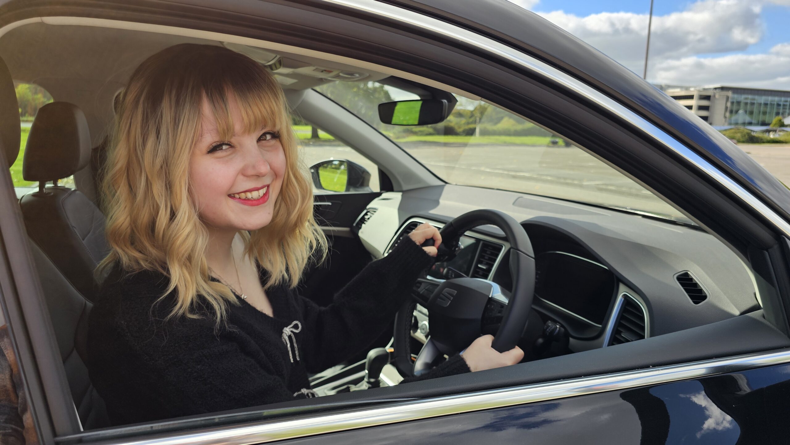 Imali, a young white woman with blonde hair, sits in a car with a black steering wheel.