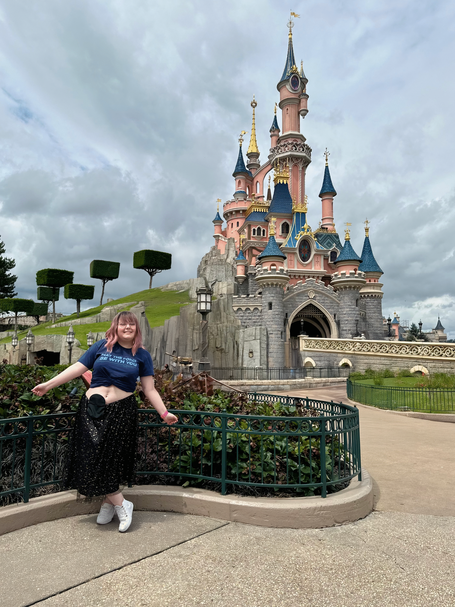A woman with pink hair, standing in front of the Disneyland Paris castle, with her stoma bag out.