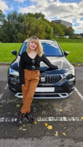 Imali, a young white woman with blonde hair, wearing a blank cardigan and burnt orange trousers, is standing with a black car.