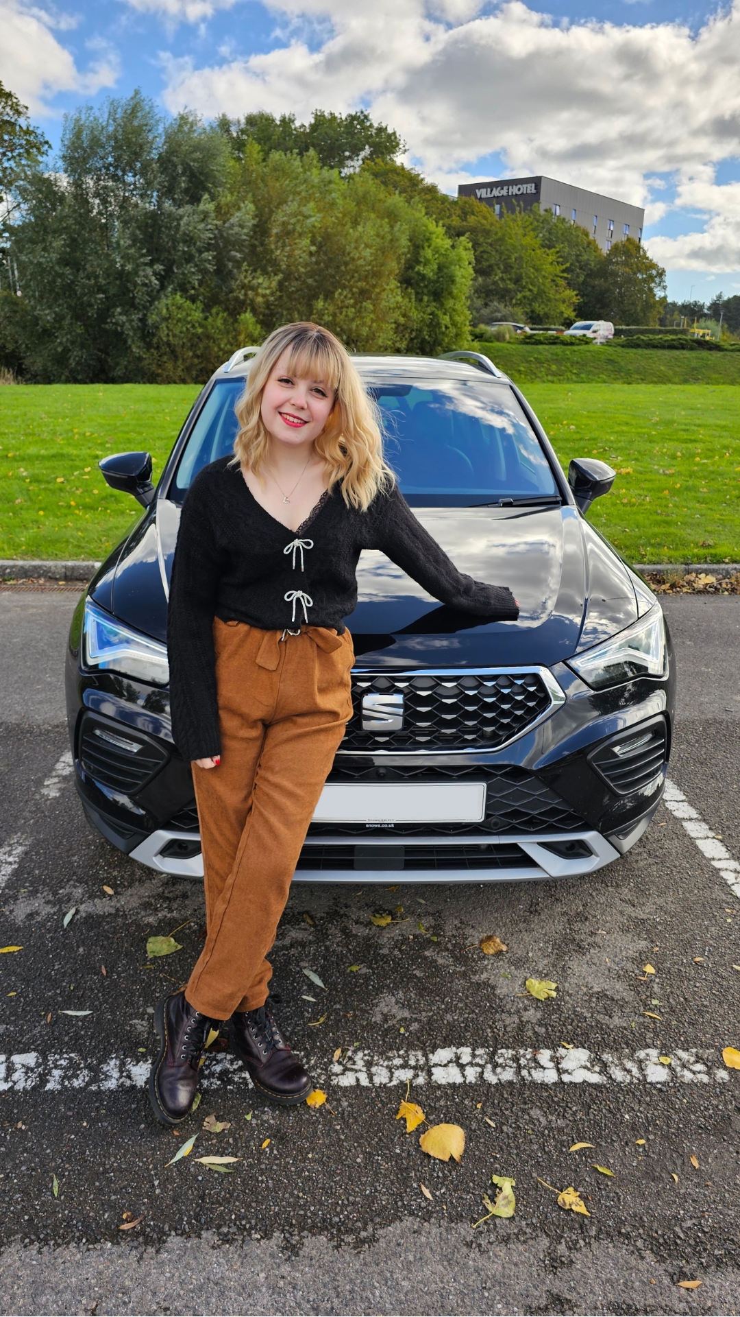 Imali, a young white woman with blonde hair, wearing a blank cardigan and burnt orange trousers, is standing with a black car.