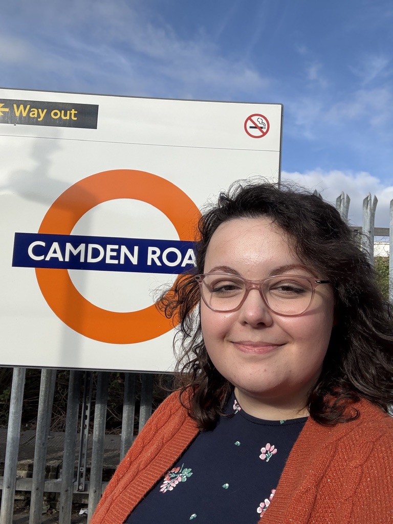A white woman, with brown curly hair. Wearing Pink glasses and an orange cardi. Sitting in front of a Camden Road tube stop sign in London. She is using a wheelchair.