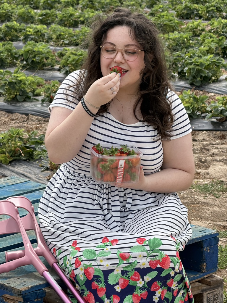 A white woman with brown, windswept wavy hair, eating a strawberry while she holds a punnet. She is wearing a white dress from Popsy London, with blue stripes, which has a navy blue band around the bottom and strawberries on it. She has paired it with red lipstick and red wedges.