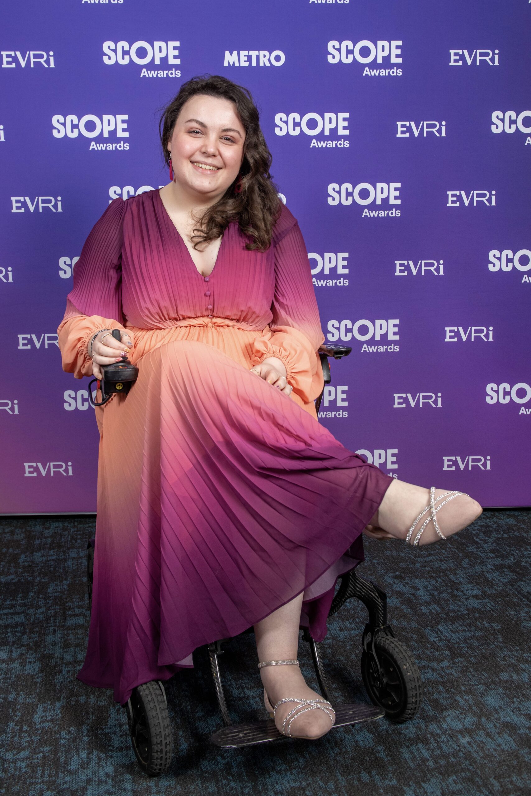 Imali, a white woman with brown curled hair, sitting cross legged in her wheelchair with a pink to orange ombre dress on and sparkly shoes.