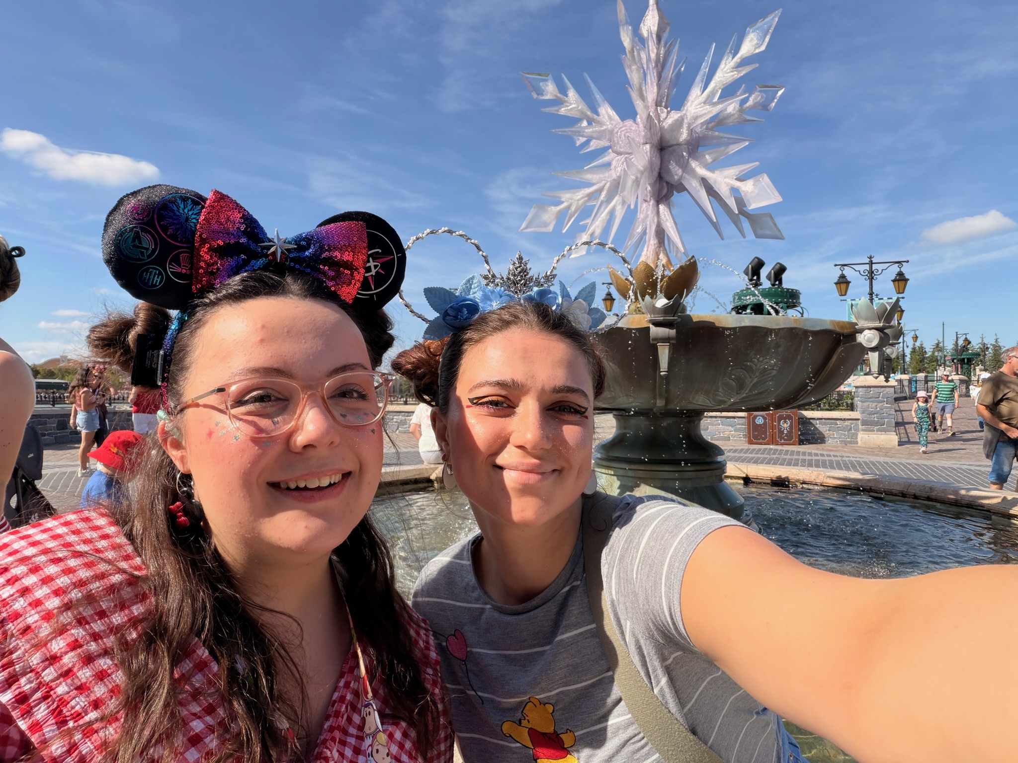 Two white women smiling at the camera, with Frozens Arendelle in the background. Theres a water feature with an icey blue snowflake in the middle. Both woman are wearing Disney ears.