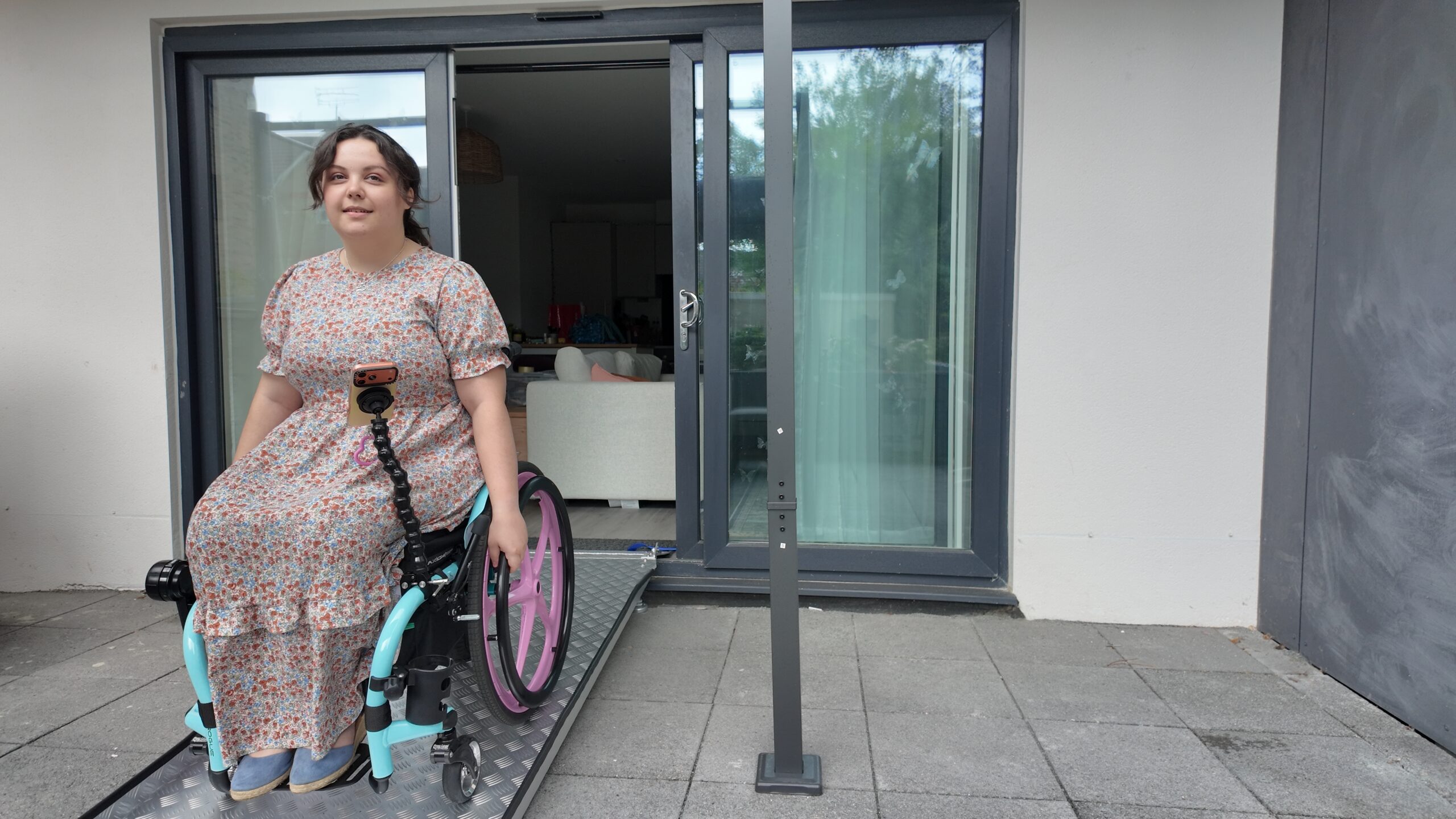 A white woman in a ditzy floral dress, sitting in a light blue wheelchair with pink wheels. She is pushing the wheelchair down a silver ramp. This post highlights the lack of accessible housing for disabled people.