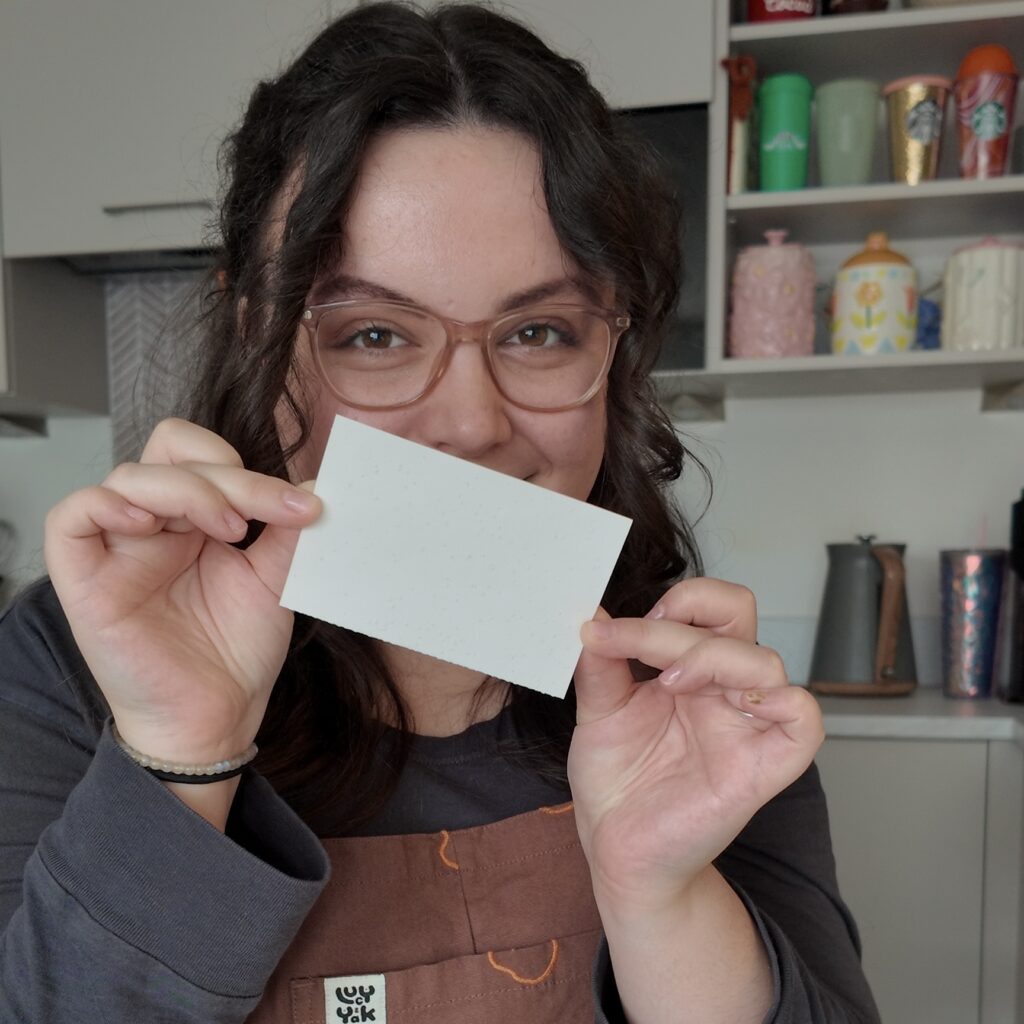 A white woman, wearing brown dungerees, holding a white rectangle laundry sheet in front of her face.