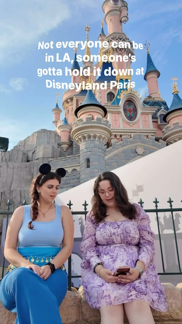 Not everyone can be in LA, someone's gotta hold it down at @disneylandparis!VD: Two white woman with brown hair, dressed up like Disney princesses, sitting on a wall with the castle in the background chatting. The clip then cuts to them meeting Aladdin and being outside the famous castle at night after the fireworks.