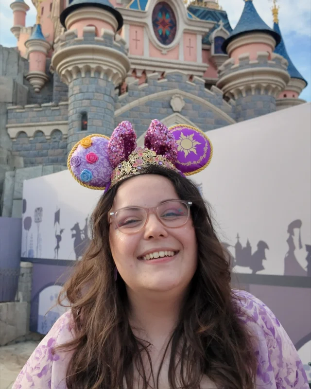 The Disney makeup of dreams... ✨ Which look is your favourite?
I truly felt like a princess every single day. 🏰VD -
Image 1: A white woman with brown wavy hair,  wearing lilac Rapunzel inspired Disney ears that sparkle. I also have a gold tiara on. I am smiling at the camera, wearing pink glasses and a lilac dress. In the background is the pink DLP castle.
Image 2: A sideways view of me, a white woman, with brown hair in a half up, half down style. I am wearing black Adventure World ears, with colourful gradients all over in sparkle. You can see my black Minnie earrings, with face glitter and hand drawn Mickey ear freckles with my pink glasses.
Image 3: A front view of the previous makeup look, now with the face glitter more obvious and my gingham red check top in view. I have taken the ears off in this selfie.
Image 4: My hair is in plaits, by my sides with orange Mickey pumpkin earrings visible. I have blue snowflake eyeshadow and a blue top.
Image 5: A close up of the silver glitter on my face and the snowflake I drew on my eyes. Pumpkin earrings in shot.